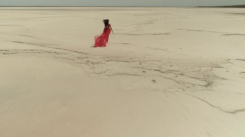 Aerial follow up young beautiful model girl woman in red scarlet flying dress runs barefoot in slow motion along dry bottom salt lake. Happiness, fashion, inspiration, beauty. Shooting slow-motion