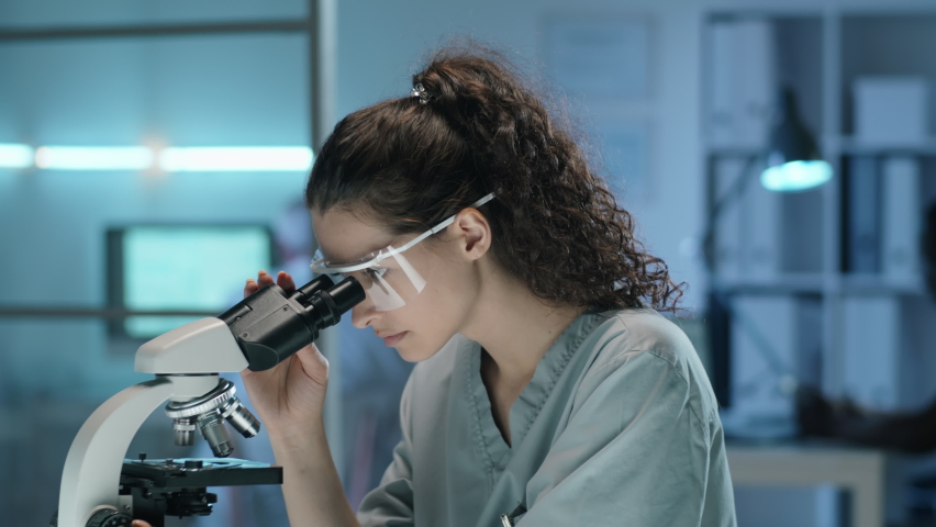 Tilt down shot of young female scientist in protective glasses and scrubs looking through microscope and taking notes on clipboard while working in lab | Shutterstock HD Video #1074706178