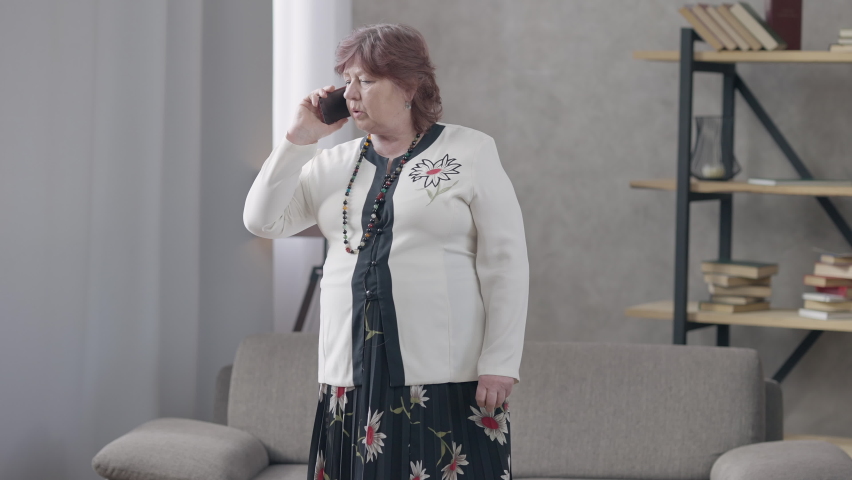 Anxious worried senior Caucasian woman talking on the phone standing in living room indoors. Portrait of stressed female retiree discussing problems with friend or relative. Lifestyle concept