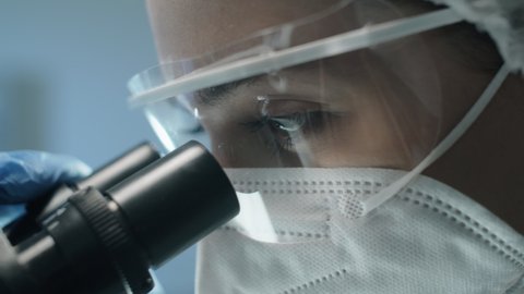 Close up shot of female scientist in protective uniform, mask and glasses looking through microscope while doing lab research during covid- outbreak