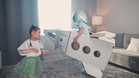 Chinese kids play in the living room of the house, a boy in an astronaut costume plays with a cardboard space shuttle, a cute girl sister runs after her brother around the room, 4k slow motion.