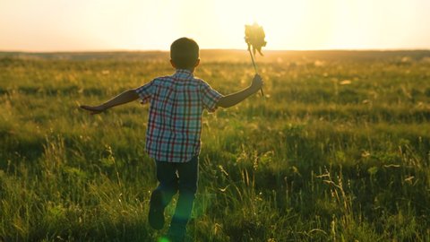 Happy kid plays with toy pinwheel outdoors in spring park in sunshine. Childhood, children. Little boy runs with toy wind turbine in his hand on summer field at sunset. Family vacation in nature.