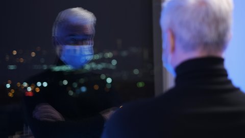 Masked businessman person in turtleneck during covid 19 looks in window. Elderly grey hair people coronavirus. White old man in facemask observe night city. Elder gray gentlemen in Covid-19 face mask.