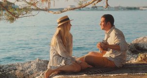 Fun attractive couple of traveler sitting on sea beach with ukulele under sunset sky in evening time. Man playing, woman listening and sing music, relaxing enjoying holidays, travel day. 4K video