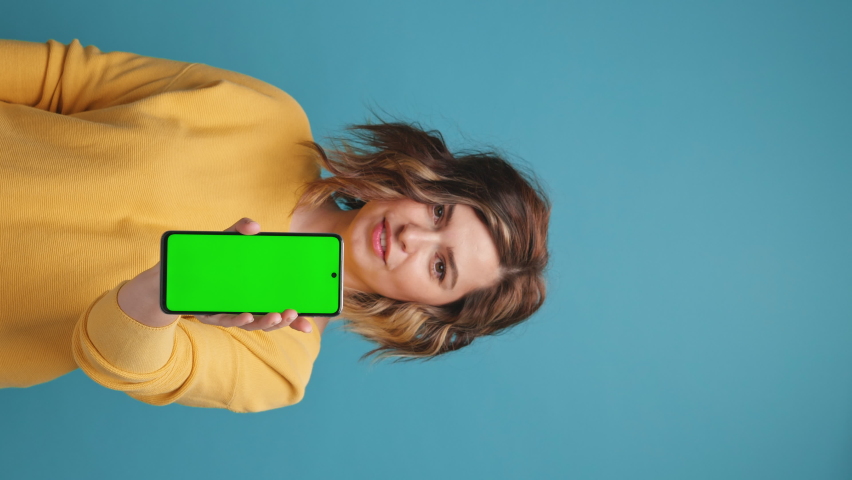 Vertical video Young woman holds smartphone with green screen index finger showing swipe gesture to copy space to advertise your products on blue background. Advertising concept promotional offers Royalty-Free Stock Footage #1074716372