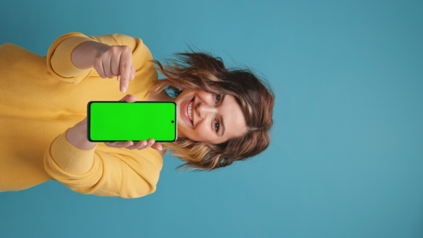 Vertical video Young woman holds smartphone with green screen index finger showing swipe gesture to copy space to advertise your products on blue background. Advertising concept promotional offers Royalty-Free Stock Footage #1074716372