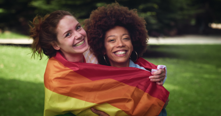 Cinematic shot of happy young girl friends of different ethnicities embracing with LGBT rainbow flag in green park. Concept of friendship, homophobia, diversity,equality, freedom, liberty, multiethnic