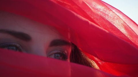 Cinematic close-up portrait face blue eyes young woman girl model in red scarf burqa hides her face. Beauty, fashion abstract film. Concubine. Red sun of desert. Shaman mystical man. Accept fate