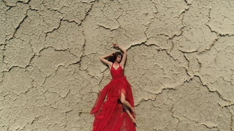Beautiful Russian girl in red dress lies asleep dreaming on cracked dry ground, scenic cracks pattern. Surreal natural landscape. Global warming. Blood comes from earth wounded planet. Aerial top down