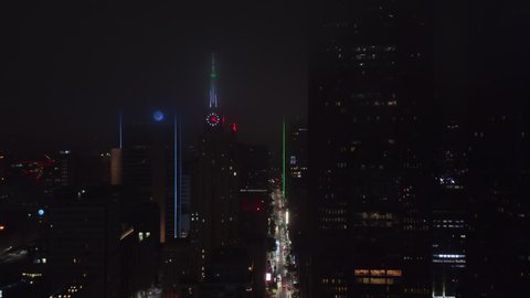 Drone flying along street between tall commercial buildings around Mercantile National Bank Building with tower and big neon clock. Night aerial view of downtown. Dallas, Texas, US in 2021