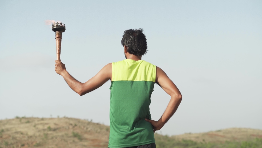 Back view of athlete proudly holding sports flame torch relay and looking mountains