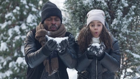 Happy carefree African American father and daughter blowing snow in slow motion laughing jumping and hugging. Relaxed man and girl having fun on winter resort. Family leisure and joy