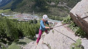 Woman hiker ascends vertical via ferrata route. She clips cables and follows metal rungs while doing so