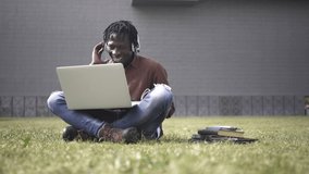 Young black African male student doing video call or webinar using laptop and headphones at outdoor university campus, sitting on the grass next to books and  notebooks