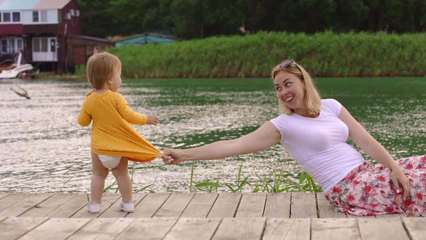 Young caucasian mother holding her little daughter on the dress and funny fallen, playing on the wooden pier by river. Side view. Slow motion. The concept of chidren