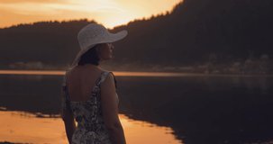 A beautiful girl in a dress and hat stands on the shore of the lake at sunset and looks around. Her back is turned to the camera. Night falls. There is a sense of peace. The girl is calm and carefree.