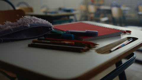 Zoom in shot of school supplies for education lying on desk. Closeup pens, pencils and notebook on wooden table. Interior of empty classroom with desks and chairs. Education concept 