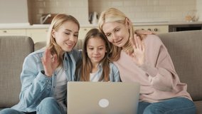Happy little girl with mother and grandmother using laptop together, sitting on couch, looking at screen, family making video call, smiling pretty child waving hand