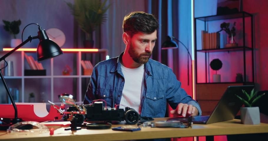 Good-looking serious thoughtful adult bearded man watching at disassembled toy car on the table and trying to repair it with screwdriwer under magnifying glass
