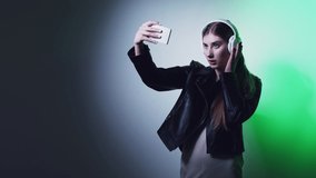 Cyberpunk people. Digital technology. Blogger lifestyle. Confident smiling girl in headphones taking selfie on mobile phone isolated on dark green neon light copy space background.