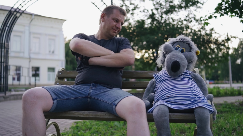 A man sits on a bench next to a stuffed wolf toy and puts his arm around it. Good mood