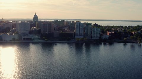 Aerial above the lake along shoreline of Madison in Wisconsin. Sunset light behind the city skyline. Domed state capitol building on the horizon