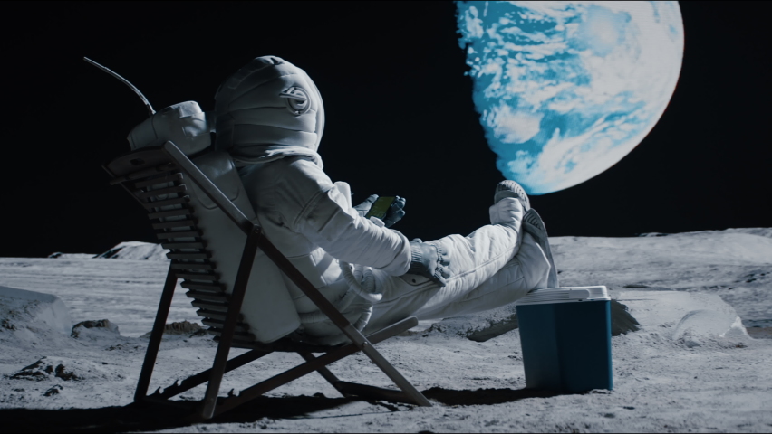 Astronaut sits in a beach chair on a Moon surface, holding phone in hands. Shot with 2x anamorphic lens Royalty-Free Stock Footage #1074778583