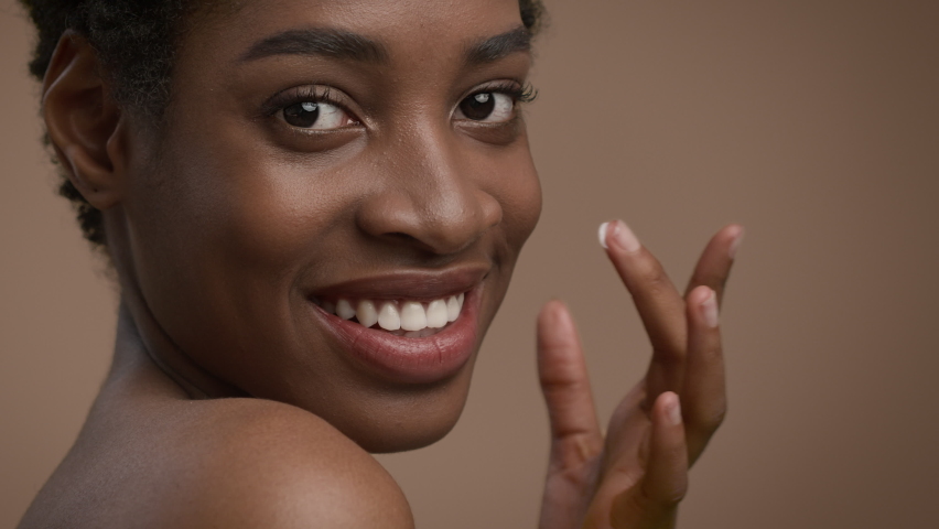 Facial Skincare. Attractive Young Black Lady Applying Cream Moisturizing Face Skin Smiling To Camera Posing Over Beige Studio Background. Natural Cosmetics, Female Beauty Routine. Cropped Royalty-Free Stock Footage #1074779846