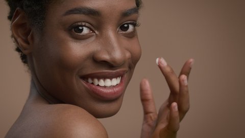 Facial Skincare. Attractive Young Black Lady Applying Cream Moisturizing Face Skin Smiling To Camera Posing Over Beige Studio Background. Natural Cosmetics, Female Beauty Routine. Cropped