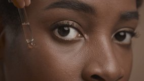 Closeup Of Black Woman Applying Facial Serum With Dropper Caring For Skin Looking At Camera Over Beige Background. Female Facial Skincare Concept. Studio Shot, Cropped