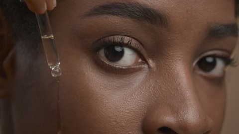 Closeup Of Black Woman Applying Facial Serum With Dropper Caring For Skin Looking At Camera Over Beige Background. Female Facial Skincare Concept. Studio Shot, Cropped