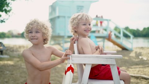 Two blond curly boys in red shorts playing on the sand beach with blue lifeguard tower, high white chair, lifeline and surfboard.