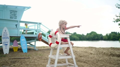 Young pretty curly boy sitting on high white chair with lifeline and posing with whistle on the beach against blue lifeguard tower. 