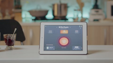 Tablet with intelligent software placed on table in kitchen with nobody in, controlling light with high tech application. Notepad with smart home app in empty house automation system