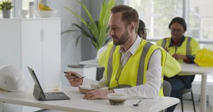 Foreman in reflective vest having video chat while eating lunch in canteen. Engineer using digital table during lunch break sitting in office and eating takeaway food