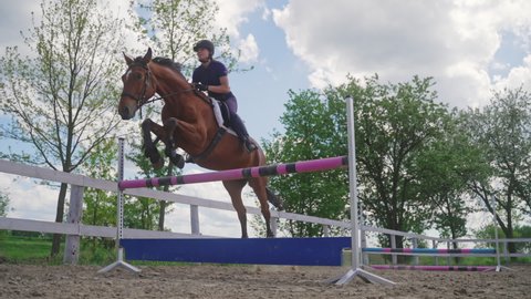 Woman rider on a horse leaping over a fence in sandy parkour. Equestrian events concept. Beautiful brown horse jumping over obstacles. Competitive woman riding a Horse. Cloudy sky in the background.