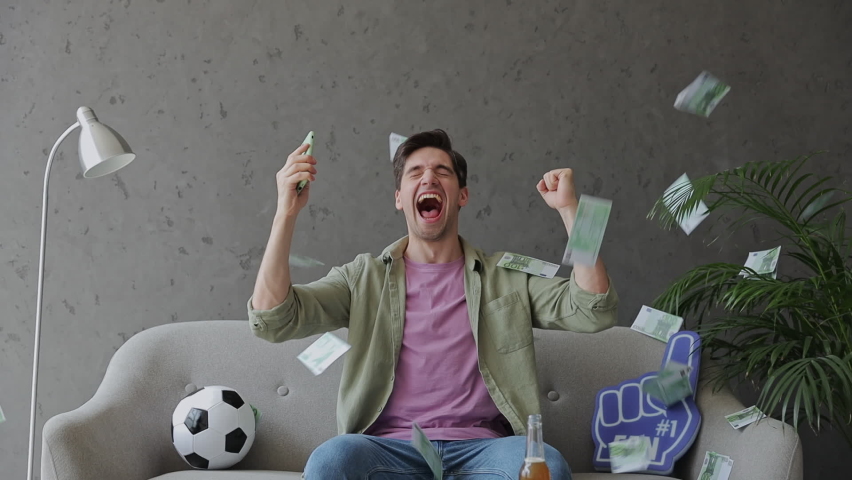 Young fun man fan in green clothes t-shirt cheer up support sport football team sit on grey sofa with soccer ball hold mobile cell phone monitors rate win fly falling money rest watch tv indoors room