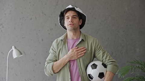 Young fun man fan in green clothes t-shirt cheer up support football team near grey sofa with soccer ball hat rest watch tv match stand sing national anthem indoors room gray color wall. Sport concept