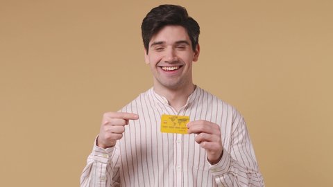Handsome stylish brunet young man 20s years old wears white striped shirt pointing index finger on mockup plastic credit bank card showing thumbs up isolated on pastel beige background studio portrait