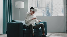 Delighted Asian guy in white virtual reality headset imagines playing djembe sitting on couch and smiling at home slow motion