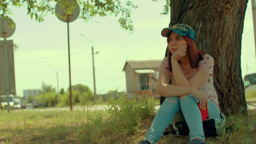 Young woman rests, sitting near tree on street. Brunette in cap looks away thoughtfully, propping chin in her hand, sitting in shade near big tree in summertime.
