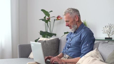 Middle-aged hipster man in smart casual shirt using laptop sitting on the couch at home, senior gray haired freelancer typing, texting, researching and exploring some information in network