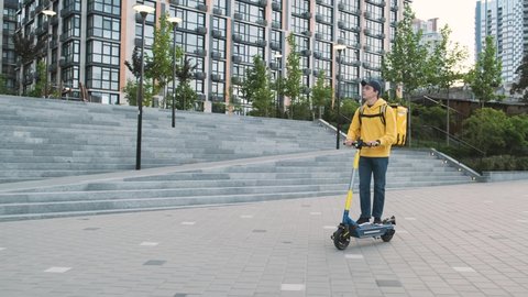 Young courier delivers package on his e-scooter early in the morning. Millenial male food courier. Deliverying goods during a pandemic