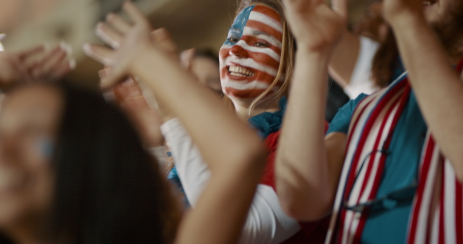 Woman with USA flag painted on her face standing in spectators and cheering for the team. Excited USA soccer supporters celebrating team success.
