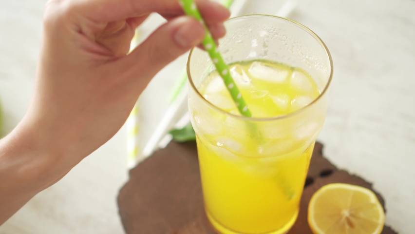 Girl without face mixes an orange drink with light green cocktail tube and drinks refreshing drink. Glass of orange soda with ice and straw on wooden slice. There are several paper tubes on white table Royalty-Free Stock Footage #1074815849