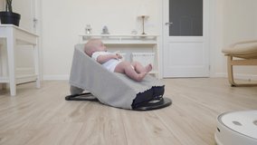 Robot vacuum cleaner performs automatic cleaning of apartment moves around baby in rocking chair, modern technology of smart cleaning. High quality 4k footage