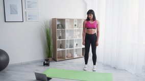 Online training. Sportive woman. Home fitness. Lockdown lifestyle. Happy athletic lady in sportswear doing warming workout on exercise mat before laptop light room interior.