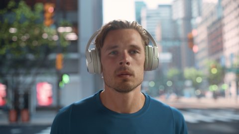 Handsome Young Man is Jogging on a Street of a Big City Center. Male is Running in Blue T-Shirt, Wearing Wireless Headphones with His Favourite Music Playing. Urban Training Workout in the Morning.