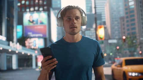 Handsome Young Man Walking on a Street of a Big City Center After Intense Fitness Workout and Using His Smartphone while Listening to Music on Hids Headphones. Urban Training Workout in the Morning.