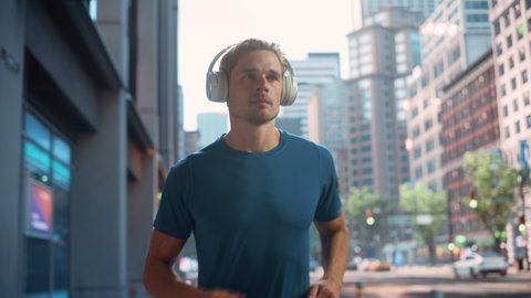 Handsome Young Man is Jogging on a Street of a Big City Center. Male is Running in Blue T-Shirt, Wearing Wireless Headphones with His Favourite Music Playing. Urban Training Workout in the Morning.
